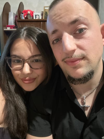 A close-up selfie shows a man and a woman posing in a room with wooden furniture and shelves in the background. The woman on the left, wearing glasses and a black shirt, has long, dark hair and is looking at the camera with a slight smile. The man on the right, also wearing a black shirt and a silver chain with a cross pendant, has a shaved head, a goatee, and is looking towards the camera with a neutral expression. The room appears to be a living space, with items on the shelves including framed pictures and decorative objects. The lighting suggests an indoor setting, possibly in a home. The image does not contain any specific indicators that would place it in Vaglen, Bulgaria.