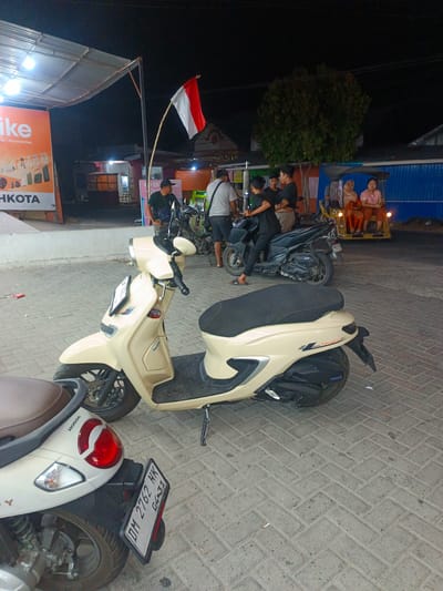 This is a nighttime photograph taken in Tenggela, Indonesia, depicting an outdoor scene with several elements. In the foreground, there are two scooters parked on a paved area. The scooter on the right is light beige and appears to be the primary subject, with its front wheel and part of its body visible. The scooter on the left is white, with its rear end and license plate clearly visible. The license plate reads "DM 2762 HK 04.31".
In the background, a group of people are gathered, some standing and some seated. A red and white Indonesian flag is prominently displayed on a pole, waving above the scene. Behind the people, there are several structures that appear to be small shops or stalls, illuminated by artificial lights. One of these structures has a large orange banner with white text that includes "like" and "HKOTA". Further in the background, a three-wheeled vehicle, commonly known as an "ojek" or "bajaj", is visible with passengers seated inside. The overall environment suggests a street or market area at night, characterized by a mix of commercial activity and public gathering.