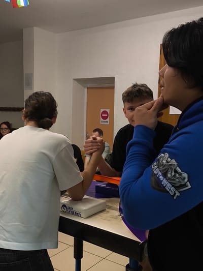 A group of people is engaged in an arm-wrestling match in what appears to be an indoor recreational setting. Two individuals, one wearing a white shirt and the other a black shirt, are actively arm-wrestling on a table designed for the sport. Their hands are clasped tightly. A person in a blue hoodie is observing the match closely. An "Arm Synergy" labeled pad is placed on the table. In the background, there is a door with a sign that says "cuisine." It can be deduced that this photograph was taken at the city of Saint-Hubert, France, due to the text "cuisine" on the sign above the door.