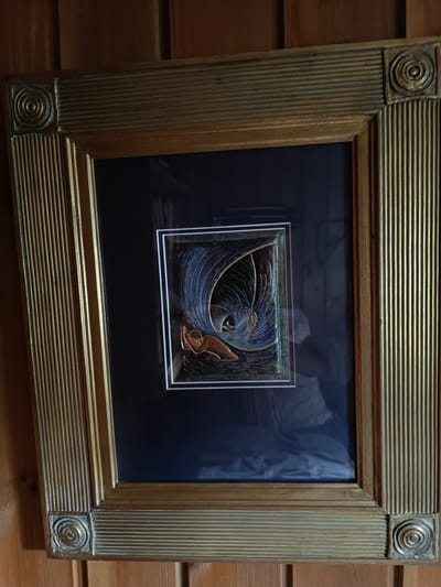 A framed artwork is displayed on a wooden wall. The artwork is rectangular and appears to be a bas-relief or embossed print. It depicts a figure, possibly a human or mythical creature, with large, fan-like wings or a flowing garment, in shades of blue, gold, and brown. The figure is reaching out or falling. The artwork is matted with a white border and framed in a wide, ornate gold frame with a textured design featuring parallel lines and circular motifs at the corners. The wooden wall behind the frame has a visible grain pattern. The lighting suggests an indoor environment, likely a home or gallery. The location is identified as Hovden, Norway.
