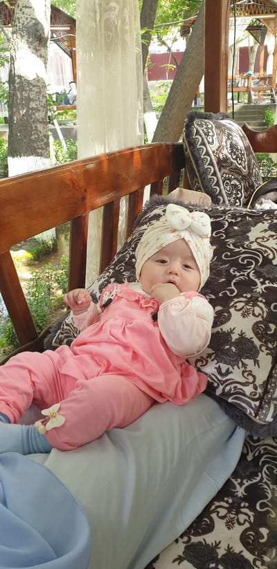 An infant, dressed in a pink outfit and a floral-patterned turban with a bow, lies on a patterned cushion on a wooden bench. The infant has their right hand near their mouth and is looking towards the camera. The bench is situated outdoors, under a canopy, with trees and greenery visible in the background. A white sheer curtain hangs to the left of the infant. The environment suggests a park or garden setting in Avval', Uzbekistan.
