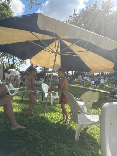 This outdoor media file depicts a sunny day at a recreational area, likely a park or community center, in Beit Yitzhak-Sha'ar Hefer, Israel. Several people are present, enjoying the warm weather. In the foreground, a young boy in red shorts is standing, and a young girl in a colorful bikini is sitting. Nearby, a man in a white t-shirt and shorts is tending to something. The scene is populated with numerous white and blue plastic chairs scattered across a grassy area, with a large, two-toned umbrella providing shade. In the background, more chairs and trees are visible, suggesting a public gathering space. The sunlight is bright, casting rays through the trees and onto the grass, indicating it's likely midday. The overall atmosphere appears relaxed and casual, with individuals enjoying an outdoor activity.