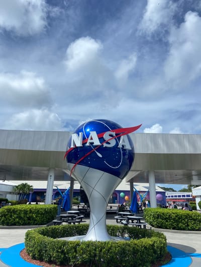 The image displays the entrance area of the Kennedy Space Center Visitor Complex on Merritt Island, Florida, United States, under a partly cloudy sky. A large, three-dimensional NASA logo sculpture, approximately 4-5 meters in height, occupies the central foreground. The sculpture features a glossy blue spherical top with the white "NASA" wordmark and a red curved stripe, supported by a silver, fluted conical base. It is centrally positioned within a circular, manicured green hedge planter, which rests on a paved surface bordered by a blue painted stripe.
Behind the sculpture, a light grey building with an extensive overhang supported by white pillars is visible, characteristic of a visitor center structure. An outdoor dining or rest area is present, furnished with multiple dark picnic tables and benches, some covered by blue umbrellas. Approximately 15 to 25 individuals are visible within this area, either seated at tables or walking. Further in the background, purple-themed structures with large, colorful spherical decorations are discernible. The upper portion of the image shows a blue sky with scattered white cumulus clouds, and several small dark specks, consistent with birds, are visible in the upper right quadrant. The scene is illuminated by natural daylight, casting moderate shadows.