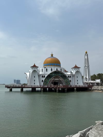 A scenic view captures the Straits Mosque, a majestic white structure with a gold-and-blue dome, sitting on stilts above calm green waters. Its unique architecture features a minaret, arched entrances with green floral patterns, and small windows. The mosque contrasts with the blue sky and the surrounding natural elements like rock formations and distant trees. A serene and spiritual scene.