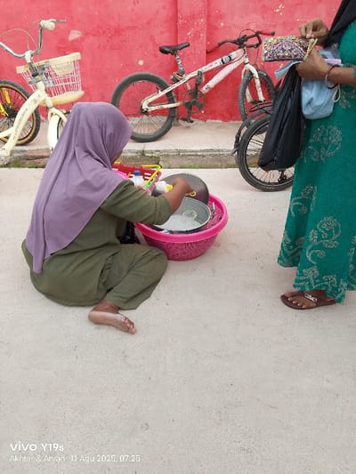 The scene is set outdoors in Jagapura Kulon, Indonesia, on a light gray concrete surface with a vibrant red wall forming the background.
Two individuals are present. On the left, a person is seated directly on the concrete ground, facing away from the viewer, clad in an olive green top and trousers, and wearing a light purple hijab. This individual is barefoot and is actively engaged in handling items within a large, bright pink plastic laundry basket. Visible items inside the basket include a dark metal pot and a clear glass lid. A smaller, red and yellow plastic basket with a white handle containing a white bottle with a yellow pump top is positioned to the left of the pink basket.
On the right, the lower torso and legs of a second individual are visible, standing. This person wears a long, teal-green dress with a darker, repetitive patterned design and brown sandals. Their hands are occupied, holding a black plastic bag, a light blue fabric bag, and a purple patterned purse with a beige strap. Bracelets are visible on their right wrist.
In the background, against the red wall, two bicycles are parked. To the left, a cream-colored "Phoenix" bicycle with a white front basket and yellow wheel accents is visible. To the right, a white BMX-style bicycle with black tires and red accents on its handlebars and frame is parked.