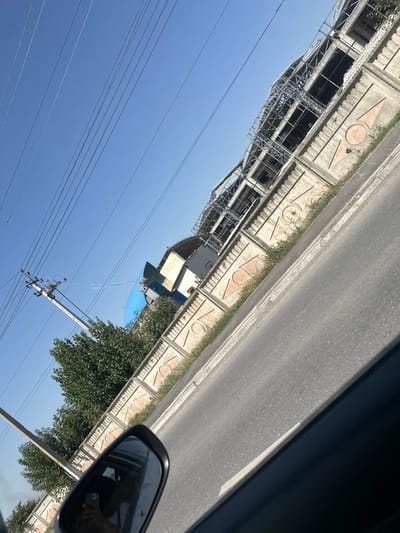 The image shows a street scene on a sunny day in Ынтымак, Kyrgyzstan. A road with a white line runs diagonally across the frame from the bottom right to the upper left. A concrete wall with decorative geometric patterns runs parallel to the road on the left side. Behind the wall, there is green foliage, trees, and several buildings, including one under construction with visible scaffolding. Power lines and a utility pole are also present in the sky. The reflection in the car's side mirror shows a person holding a smartphone. The overall environment appears to be a suburban or semi-urban area.