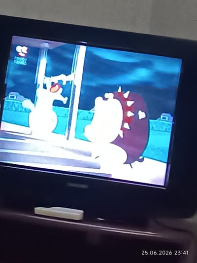 The image is a photograph of a television screen displaying a cartoon. The cartoon appears to be a scene from a video game or animated show featuring two characters. On the left is a white, anthropomorphic dog-like character with large ears and an apple in its mouth. On the right is a round, purple character with spikes around its head and a red collar. The background is a blue, stylized landscape with hints of buildings. The scene is lit as if it is nighttime or indoors with artificial lighting. A timestamp "25.06.2026 23:41" is visible in the bottom right corner of the television screen, indicating the date and time the photograph was taken. The television is placed on a dark surface, likely furniture. A small white object, possibly a remote control or a small electronic device, is visible on the surface in front of the television. The overall impression is a casual scene of someone watching television.