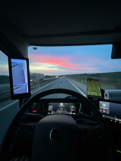 The image presents a driver's perspective from within the cabin of a Volvo heavy goods vehicle (truck), identified by the emblem on the steering wheel, traveling on a multi-lane asphalt highway in Wallhausen, Germany. The exterior scene depicts dusk or dawn, characterized by a sky with blue hues transitioning to prominent horizontal bands of pink and orange clouds. The highway appears to have light traffic, with several distant vehicles, including other trucks. Green verges and distant trees line both sides of the road.

Within the dimly lit cabin, the steering wheel is central. Directly beyond it, a digital instrument cluster displays a speed of "82" (likely km/h), the time "15:45," and various vehicle operational data including gear selection and fuel levels. To the left of the driver, a vertical digital monitor functions as a virtual side mirror, showing a real-time feed of the highway and a truck in the left lane. To the right, a smartphone or dedicated GPS device is mounted, displaying a navigation map with a road network. Further to the right, a larger infotainment screen is visible, displaying "DeutschlanRadio" and media controls. No individuals are directly visible, as the image is taken from the driver's viewpoint.