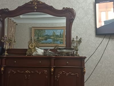 The media file shows a domestic scene in Izberbash, Russia. A vintage wooden dresser with ornate gold detailing serves as the focal point, topped with various decorative objects. A large, ornate mirror hangs above the dresser, reflecting a framed landscape painting of a serene lake surrounded by trees. On the dresser, a golden clock with intricate carvings sits next to a white fabric object, possibly a cushion or folded cloth. To the right of these items, a small grey tabby cat is resting, with its head tilted and eyes partially closed. Further to the right, on the dresser's surface, is a decorative glass bowl with a metal stand, and a brass candelabra. To the far left of the dresser, another brass candelabra is visible. The wall behind the dresser is covered in textured wallpaper with a floral pattern. To the right of the dresser, a flat-screen television is mounted on the wall, displaying a still image of a movie scene with Russian text at the top: "Зелёный фонарь (2011)". The lighting suggests it could be daytime, possibly indoors with artificial lighting. There are no visible weather cues or strong emotional indicators beyond the relaxed posture of the cat.