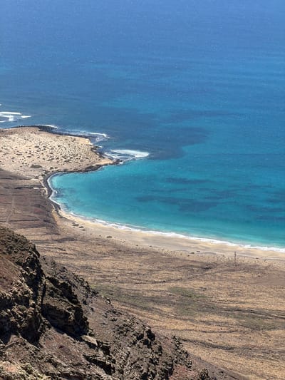 This image shows an aerial view of a beach and surrounding landscape. The location is in Haría, Spain. The ocean is blue and deepens in color further away from the shore. A sandy beach curves along the coastline, transitioning into a rugged, brown landscape with sparse vegetation. A rocky cliff forms the edge of the land, its texture rough and uneven. People are visible on the beach. The perspective is from above, looking down on the scene.