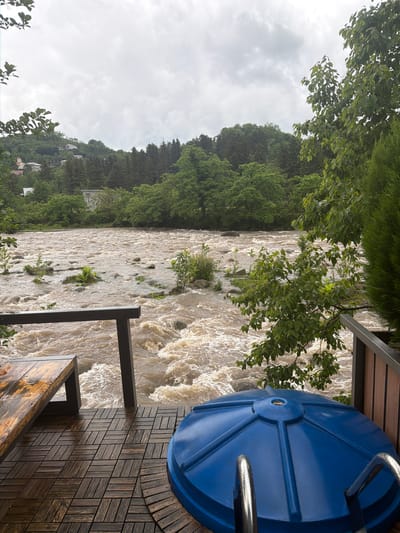 From a vantage point in Kapreshumi, Georgia, a wide, turbulent river flows, swollen with muddy water. The river's strong current is evident, with visible rapids and froth. Lush green trees line the banks, indicating a densely vegetated area. The image is taken from a wooden deck with a blue, round object, possibly a hot tub cover. A section of railing is visible. The weather appears overcast. Houses are visible on the hill in the background.
