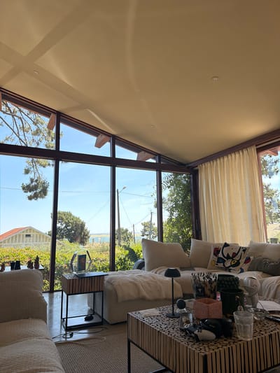 The image depicts a modern living room with large windows offering a view of the landscape and sea in Lège-Cap-Ferret, France. The room features a large sectional sofa with throw pillows, a coffee table adorned with various objects including video game controllers and decorative items, and a side table with a lamp and a hexagonal frame. The sunlight streams in, casting shadows on the ceiling and the floor. The exterior view shows greenery, houses, and a body of water, suggesting a coastal or lakeside setting. There are no people visible in the image. The overall atmosphere is one of relaxed, contemporary living.
