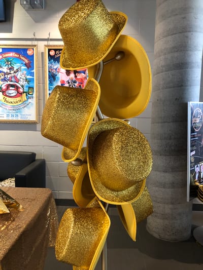 The image depicts a display of glittery gold hats in what appears to be an indoor venue, potentially a theatre or event space. The hats are arranged on a rack, with some tilted and others hanging. In the background, two posters are visible, one on the left with illustrations of characters and text in Russian, and another to its right also featuring illustrations and text, possibly related to a performance or event. A large, textured concrete column stands to the right of the hats. A table covered in a gold sequined tablecloth is partially visible in the lower left corner. A black leather couch is seen behind the table. To the far right, a vertical sign with "star school" branding is present, with additional text and images below it. The overall lighting suggests an interior environment. There are no people visible in the image. Based on visual cues, this scene is likely located at a venue in the city of Vaughan, Canada, such as the Woodbridge College or a similar performing arts or community center that hosts events and may utilize such thematic decorations.