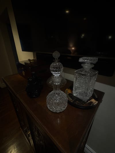 An interior scene in Miami Shores, United States of America, depicts a dark wooden credenza positioned against a wall, below a large, dark television screen. The credenza, exhibiting carved details, supports multiple objects. Centrally, a clear, faceted crystal decanter with a spherical body and a matching stopper stands prominently. To its right, atop a circular tray with dark and light mosaic elements, is a taller, clear rectangular decanter featuring a floral or leaf pattern and a rectangular stopper. To the left of the central decanter, a dark, opaque lidded jar or container is present. Further back on the credenza, behind the opaque container, is a small brass bird figurine with outstretched wings. A third, partially obscured clear glass decanter, appearing to have a fluted base, is situated behind the central decanter. The television screen in the background reflects several indistinct bright light sources. A light-colored wall is visible to the left of the television. The floor, visible in the lower-left, is dark wood. No individuals, actions, or interactions are visible.