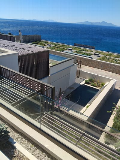 The image shows a high-angle, slightly rotated view of modern architecture near a body of water, possibly the coast. The body of water appears to be a deep blue, possibly the sea, with land faintly visible in the distance.

The architecture consists of structures with horizontal slats and glass barriers that suggests balconies. The color palette is primarily shades of white, beige, and gray, with glimpses of dark blue water in swimming pools.

The surrounding area is adorned with greenery, including shrubs and grass, which provide contrast with the hard lines of the buildings. The scene appears to be taken from an elevated vantage point, such as a higher level of the structure. The location in Pyli, Greece, cannot be definitively confirmed solely from the visual information.