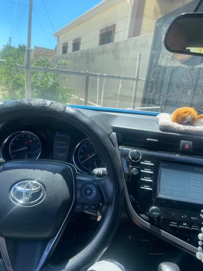 The image shows the interior of a car, looking out at a street scene. The dashboard is visible, with the steering wheel, instrument cluster, and infotainment system. A small plush toy animal is placed on top of the dashboard. Outside the car, a building and a gate are visible, along with trees and a power line. The sky is blue and clear. The car appears to be parked on a street in a residential area. The location is identified as Рахат ауылдық әкімдігі, Kazakhstan.