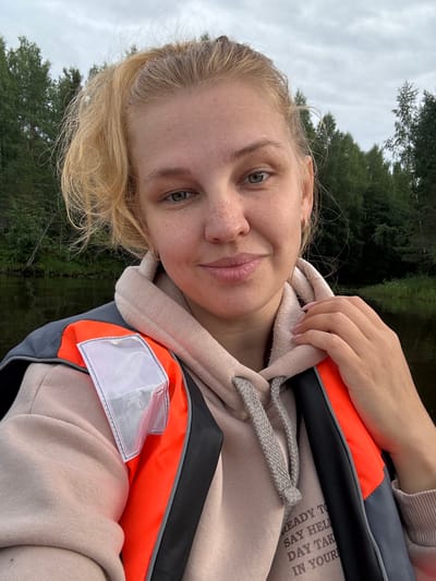 A young woman with blonde hair tied back in a ponytail and freckles on her face smiles for a selfie. She is wearing a light pink hooded sweatshirt and a bright orange and gray life vest. She has her hand resting on her shoulder. The background shows a body of water and a dense forest with a cloudy sky. The location is specified as Anteplevo (Okhotinskoe S/P), Russia.