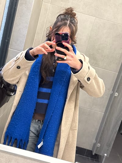 A woman in a tan coat takes a mirror selfie. She wears a blue, fringed scarf over a striped sweater and jeans. Her hair is in a messy bun. She holds a smartphone with a multicolored case in front of her face. Her fingers display several rings. The background includes tiled walls and a glimpse of a purse.