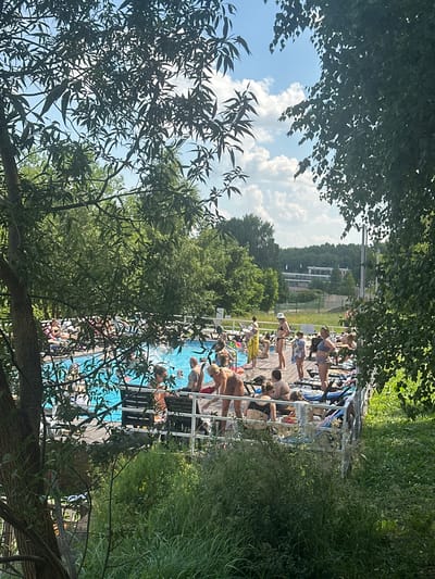 This image depicts a sunny day at an outdoor swimming pool complex, likely in Dal'nie Prudishchi, Russia. The scene is filled with people enjoying leisure activities around a vibrant blue pool. A wooden deck surrounds the pool, furnished with lounge chairs and towels, indicating a relaxed atmosphere. People are seen sunbathing, chatting, and interacting, suggesting a social gathering or a day out with family and friends.
The natural environment plays a significant role, with lush green trees and grass framing the pool area. The foliage, particularly the leaves in the foreground and scattered throughout the scene, suggests it's either spring or summer. Sunlight filters through the leaves, casting dappled shadows and highlighting the bright blue of the water. The sky is partly cloudy, with white cumulus clouds against a clear blue backdrop, indicating fair weather.
The perspective of the image, taken from slightly above and looking down, captures a bustling scene. In the background, there are some buildings nestled amidst a wooded hillside, offering a glimpse of the surrounding landscape and hinting at the location's setting. There are no visible text elements in the image. The overall mood appears cheerful and relaxed, typical of a pleasant day at a recreational facility.