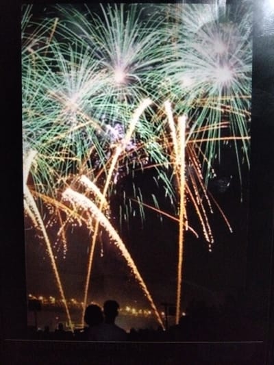 The image depicts a nighttime scene featuring fireworks illuminating the sky over a cityscape. Several large, fan-shaped fireworks with green and white light streaks dominate the upper portion of the frame. Below these, orange, glowing trails of light descend from bursts of fireworks, some appearing as thin lines and others as wider streaks with smaller dots of light. In the foreground, silhouetted figures of people are visible, appearing to watch the fireworks display. Two figures in the middle ground are more defined, with one appearing to have longer hair. A faint outline of buildings and lights from the city can be seen in the distance, beneath the fireworks. The overall environment is dark, indicating the display is taking place at night. There is no specific indication of the location being Kualaraja, Indonesia, within the image itself.