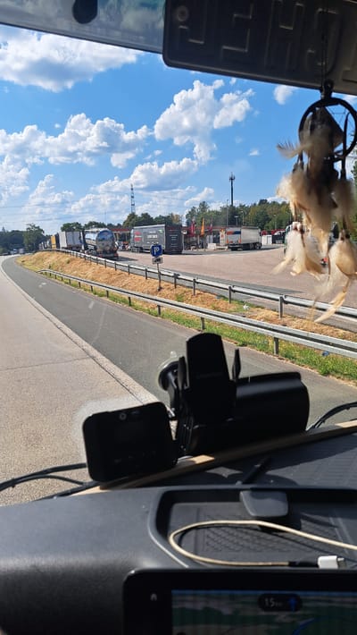 The image shows a view from inside a vehicle, likely a truck, looking out at a highway rest stop in Siegen, Germany. Several trucks are parked in a line at the rest stop, some of which are tanker trucks. The sky is blue with fluffy white clouds. A dreamcatcher hangs from the rearview mirror of the vehicle, obscuring a small portion of the view. There are trees and vegetation surrounding the rest stop. In the foreground, inside the vehicle, a dash camera and a mobile phone mount are visible. A GPS screen is also visible at the bottom of the frame.