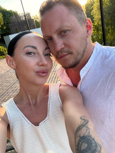 A photograph depicts a man and a woman posing closely together, likely for a selfie. The man has fair skin, short blond hair, a beard, and a tattoo on his left arm. He is wearing a white shirt. The woman has fair skin, dark hair pulled back, and is wearing a white crocheted top with a necklace. They appear to be standing outside with a view of trees and a wooden structure in the background. Based on the structure, the location is potentially Krasny Klyuch, Russia. The image is taken at an angle, looking slightly up at the subjects.