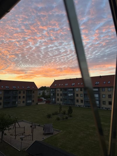 The image depicts a view of a courtyard between apartment buildings in Roskilde, Denmark, during a sunset. The sky is filled with scattered clouds that are tinged with orange and pink hues, indicating the setting sun. In the foreground, a playground area is visible with sand, wooden climbing structures, and a small slide. Beyond the playground are two multi-story apartment buildings with red tile roofs. The buildings are constructed with beige bricks and feature balconies and numerous windows. A patch of well-maintained green grass surrounds the playground and extends between the buildings. In the background, trees and other buildings can be seen, as well as a white van. The perspective is from an elevated position, looking down into the courtyard, and a metal structure, possibly part of a balcony railing or window frame, partially obstructs the view on the right side of the image. There are no people visible in the scene.