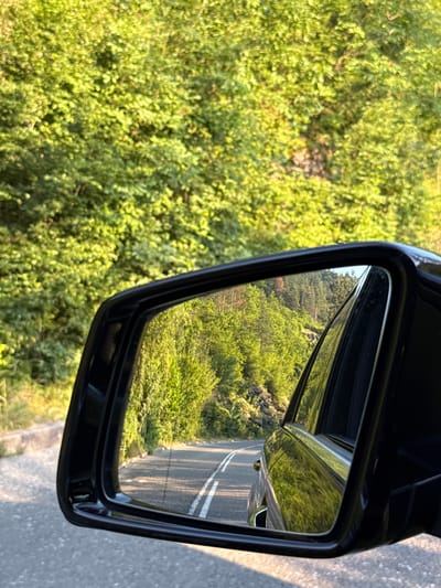 The image shows a close-up of a car's side mirror, reflecting a rural landscape. The mirror itself is black and framed in black plastic, attached to the car door. The reflection in the mirror captures a road with double white lines, indicating it is a two-lane road. To the left of the road, there is a dense green hillside covered in lush vegetation, with patches of sunlight filtering through. In the background of the reflection, more green hills can be seen, suggesting a mountainous or hilly terrain.
Outside the mirror, to the left of the frame, the actual scene is predominantly green foliage, likely bushes or trees, bathed in what appears to be late afternoon sunlight given the warm glow and the shadows cast. The road surface is visible on the right side of the image, also catching the sunlight, with its texture of asphalt and the white lines clearly defined. The scene evokes a sense of travel through a natural, verdant environment. There is no visible text in the image. The time of day is likely late afternoon or early evening due to the quality of light. The location cues of lush greenery and a rural road suggest a setting in a place like Taran, Bulgaria, known for its natural beauty.
