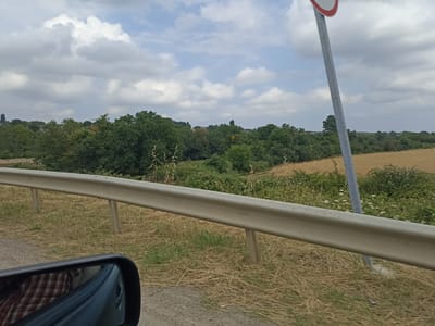 The image shows a roadside view with a guardrail in the foreground. Beyond the guardrail, there is dry grass and then a field of golden crops, likely wheat or barley. Further in the distance, there is a dense line of green trees, and beyond that, the sky is partly cloudy with a mix of white and gray clouds. A metal pole with a circular sign, predominantly white with a red border, is visible on the right side of the image. The reflection in the side mirror on the left shows a person wearing a red and white checkered shirt. There is no specific information about Xucbala, Azerbaijan, in the image.