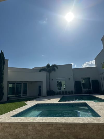 The image depicts a modern residential backyard in McAllen, United States of America, under a bright, clear blue sky with minimal cloud cover and prominent direct sunlight causing lens flare in the upper right quadrant. The foreground features a raised rectangular swimming pool, likely a spa, exhibiting a stacked light-colored stone facade and light blue tiled interior, connected to a larger rectangular pool. Both pools contain turquoise water and are surrounded by light-colored stone paving. To the left of the pools, a manicured green lawn is visible. The background consists of a multi-sectioned, light-colored modern house with flat roofs. The house features several rectangular windows; one on the right has a partially raised exterior rolling shutter, and a covered patio area on the left includes large sliding glass doors and a visible ceiling fan. Vegetation includes a tall, slender cypress-like tree on the far left and a palm tree, from which a lightning rod extends, positioned in the mid-ground. Dark green shrubbery lines the base of the house structure. No individuals are present in the scene.