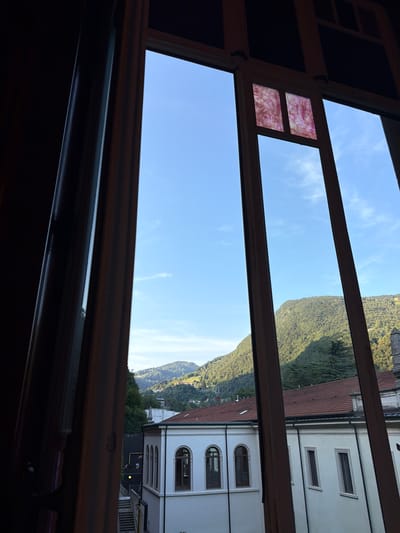 This media file depicts a view from a window in San Pellegrino Terme, Italy. The scene is dominated by a clear blue sky with sparse white clouds, suggesting fair weather during the daytime. The window frame, made of dark wood, divides the view into several sections.

In the upper left portion of the window, a small pane of decorative stained glass with swirling pink and white patterns is visible. The main view through the window showcases a lush green mountainside, characteristic of the Italian Alps, dotted with trees and some buildings nestled into the slope. The lower part of the view reveals buildings with white facades and tiled roofs. One building features arched windows, typical of older European architecture. A yellow circular object, possibly a light fixture or sign, is visible on one of the buildings. The overall impression is a peaceful, scenic landscape bathed in natural light.

No people or specific activities are discernible in the image. The composition, looking outwards from an interior space, evokes a sense of quiet observation or contemplation. There is no visible text in the image.