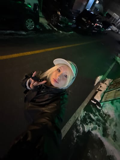 An overhead-angle night photograph captures a blonde individual in Namyangju-si, South Korea. The subject, positioned centrally in the lower half of the frame, wears a white baseball cap with a green underside, a black leather jacket, and a light-colored hooded garment. They are looking directly at the camera, holding a device for a selfie, with their right hand raised, fingers forming a 'V' gesture. The background consists of a dark asphalt surface with a prominent yellow road line and a faded white line. Patches of snow are visible on the ground along the right side and near several parked dark-colored vehicles in the distance. Green ambient lighting is present, particularly illuminating the snow and parts of the background.