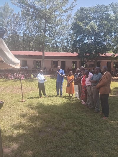 The image displays an outdoor scene in Idakho South ward, Kenya, under a clear sky. A grassy expanse occupies the foreground. In the mid-ground, a group of eight adults is positioned. One male, wearing a blue polo shirt and light-colored trousers, stands centrally, holding a microphone to his mouth, appearing to speak. To his right, six individuals (five males, one female) are lined up, facing left towards the speaker. Their attire ranges from casual to semi-formal. To the speaker's immediate left, another male in a light blue shirt and dark trousers stands facing the speaker, with his hands clasped. On the far left of the frame, a segment of a light-colored canopy or tent structure is visible, supported by a pole. In the background, behind the canopy, a large group of individuals, predominantly children, are seated in rows on the ground. Further back, a single-story building with light-colored walls and a reddish corrugated iron roof is observed, featuring multiple windows and doorways. Several individuals are visible seated in the openings of this building. The background is dominated by numerous trees, including tall, slender specimens and broader-canopied varieties.