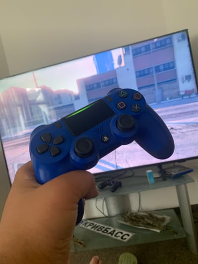 A person's hand is holding a blue video game controller in front of a television screen displaying a video game scene. The television is mounted on a wall. A glass table is visible below the television, with a video game console, a small blue object, and a stack of papers or books on it. A license plate with Cyrillic text is also visible on the table. The scene appears to be indoors. The image is taken from a low angle, looking up at the controller and television. The lighting is dim, with the television screen being the primary light source. The overall impression is a casual setting, likely a living room, where someone is playing a video game. There is no indication of the specific city of Vejprty, Czechia.