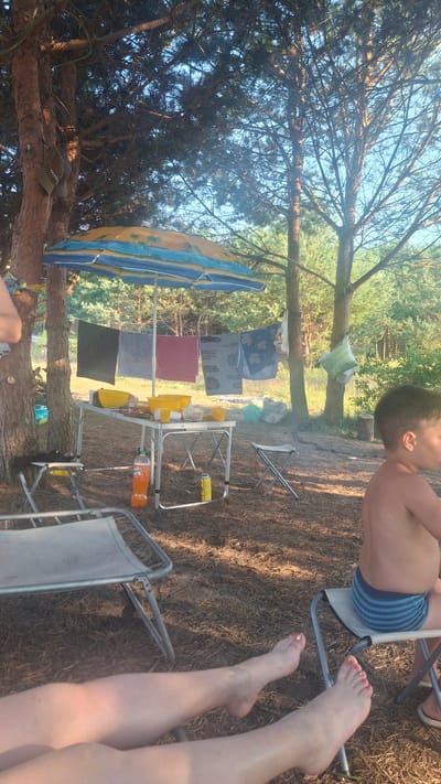 The image depicts a scene set in a wooded area, likely a campsite, in Навасвержанскі сельскі Савет, Belarus. The foreground features the legs of a person or people relaxing on chairs, with their feet visible. To the right, a young boy is seated on a small folding chair, facing away from the camera.
In the midground, a portable table is set up under a beach umbrella adorned with a tropical scene. Various yellow bowls and cups are arranged on the table, suggesting a meal or gathering. Several towels are hung on a clothesline strung beneath the umbrella. A bottle of orange soda and a yellow can are also visible on or near the table. Two small folding stools are placed around the table, and a larger portable chair is situated to the left.
The background consists of a dense forest with tall trees and undergrowth. The ground is covered with pine needles and dry leaves. The lighting suggests it is daytime, with sunlight filtering through the trees. There are a few plastic bags and a green, possibly helium-filled, balloon hanging from a tree in the distance. The overall impression is a relaxed outdoor activity in a natural environment.