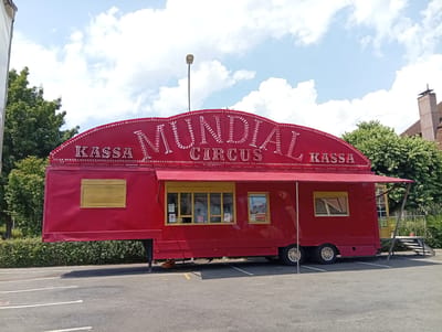The image shows a bright red circus trailer with the words "MUNDIAL CIRCUS" emblazoned in white, sparkly letters on its curved roof. The trailer has a large red awning extending over a windowed section, which appears to be a ticket booth or concession stand. Yellow blinds cover one window on the left side of the trailer. The trailer is parked on an asphalt surface with white painted lines, suggesting a parking lot or open area. Behind the trailer, there are dense green trees and bushes, and in the background, a building with a chimney is visible. The sky is a mix of blue with scattered white clouds. A street lamp stands tall behind the trailer. The overall scene depicts a mobile circus setup, likely in an outdoor location within the city of Langenthal, Switzerland, possibly during a fair or event. There are no people visible in the image.
