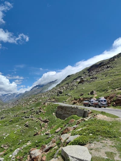 The image depicts a mountainous landscape in Koksar, India, under a bright blue sky with scattered white clouds. A winding asphalt road curves through the scene, bordered by steep, green, rocky slopes. Several trucks, likely carrying water or fuel, are visible on the road. The terrain is characterized by lush green grass interspersed with numerous rocks and boulders of varying sizes. In the distance, more mountains are visible, some with snow-capped peaks and shrouded in mist. The overall impression is one of a remote, high-altitude region with dramatic natural beauty.