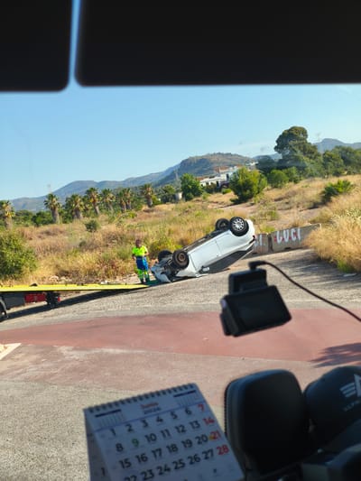 A white car is flipped on its roof on a roadside next to a tow truck. A worker in a neon yellow vest and blue pants is standing next to the car, appearing to be supervising the scene. The tow truck is yellow and is positioned behind the overturned car. There are concrete barriers with graffiti on them near the car. The background shows a dry, grassy hillside with palm trees and mountains in the distance. The scene is illuminated by bright sunlight. A calendar showing June 2024 is visible in the foreground, suggesting the image was taken during that month. A dashcam is also mounted on the windshield in the foreground. The location is on a rural road in Nàquera, Valencia, Spain.
