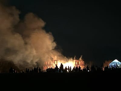 At night, a large wooden structure resembling a building burns intensely, engulfed in flames and thick smoke. Many silhouetted figures stand watching from a distance, situated on a slight rise. To the right, a white tent is partially visible. The sky is dark and clear, emphasizing the fire's brightness.