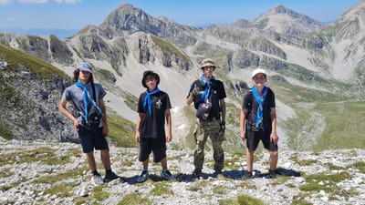 The image shows four young males standing on a rocky, mountainous terrain under a clear blue sky. The individuals are wearing casual clothing, with the exception of blue and yellow scout scarves around their necks. Each person is equipped with a backpack, suggesting an outdoor excursion or hiking trip. In the background, a vast mountain range stretches across the horizon, characterized by rugged peaks, slopes covered with sparse vegetation, and rocky scree. The lighting indicates it is daytime, likely midday, given the bright sunlight casting shadows. The scene appears to be a remote, natural landscape, potentially within the Gran Sasso mountain range, which is near the city of L'Aquila in Italy.