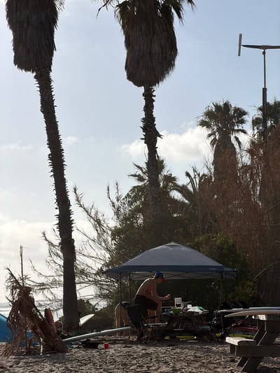This image depicts a beach scene in San Clemente, California, under a clear, bright sky with scattered clouds. In the foreground, a person is seen sitting at a picnic table under a pop-up canopy. They are shirtless, wearing dark shorts and a blue baseball cap, and appear to be holding food. Several surfboards are visible, some leaning against the table and others lying on the sand. Other picnic tables and chairs are also present, along with various beach gear. In the background, palm trees and dense vegetation line the shore, with the ocean visible in the distance. The overall atmosphere suggests a casual, recreational gathering on a sunny day.