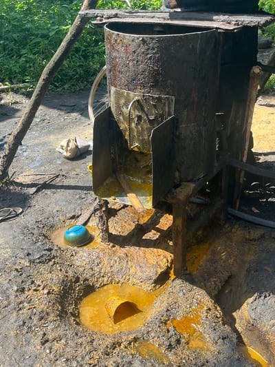 The image displays an outdoor, makeshift industrial scene bathed in bright daylight, consistent with the location context of Aghalokpe, Nigeria. The central subject is a large, dark, and heavily grimy metal cylinder or drum, likely a processing tank, supported by a rusty, rugged metal framework and integrated wooden beams. This setup suggests a small-scale, possibly informal, industrial operation.

The ground around the equipment is a mixture of dark soil and gravel, extensively stained and pooled with a viscous, striking yellow-orange liquid. This substance coats the metal structures and forms puddles, indicating the processing or handling of materials such as crude oil, chemicals, or other mineral-rich substances. One puddle on the right contains a small, bright blue bowl-shaped object, while another holds a smaller, open metal pipe or container also filled with the yellow liquid.

Hints of human presence include a partially visible bicycle wheel behind the large cylinder, a crumpled light-colored bag on the ground, and a section of a flip-flop sandal near the blue object. Dense green foliage in the background signifies a rural or semi-rural environment. The scene implies an ongoing industrial activity, possibly involving distillation or refining, carried out with rudimentary equipment in potentially hazardous conditions. No visible text is present in the image.