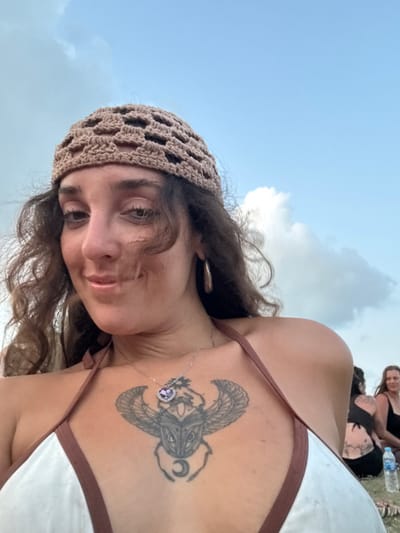The image presents a close-up outdoor scene in Ban Pang Pring, Thailand. A female individual with a medium skin tone and long, wavy dark brown hair occupies the foreground, looking directly at the camera with a subtle upward curve of the lips. She wears a light brown crocheted open-weave skull cap, a white bikini top with brown trim, and a silver necklace featuring a heart-shaped purple stone pendant encircled by smaller clear stones. A silver-colored teardrop hoop earring is visible on her right ear. A prominent scarab beetle tattoo with outstretched wings and a crescent moon design is visible on her upper sternum. The background features a blue sky with scattered white cumulus clouds. To the far right, two other individuals are partially visible in the blurred mid-ground: one from behind with a lower back tattoo, and another with a dark top, facing forward with a faint smile. A clear plastic water bottle with a blue cap rests on the ground near the background figures.