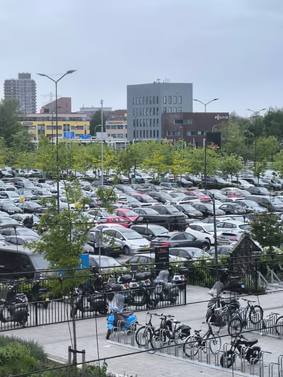 The image shows an extensive parking lot in Leidschendam, Netherlands, filled with numerous cars of various makes and colors. A bicycle and scooter parking area is visible in the foreground, with several bicycles and scooters parked. In the background, buildings are visible, including a modern gray building, a yellow and blue building, and a brown building labeled "Politie." Streetlights line the parking area. The sky is overcast.