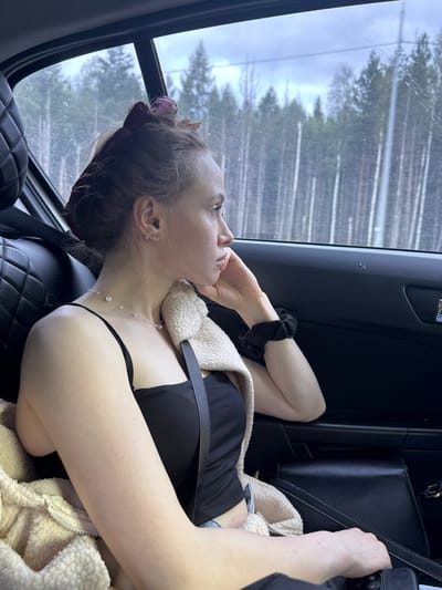 A young woman with brown hair pulled back is sitting in the back seat of a car, looking out the window at a forest. She is wearing a black tank top, a pearl necklace, and a tan fuzzy jacket draped over her shoulders. Her left hand is resting on her cheek, and she has a black scrunchie on her right wrist. The car interior is visible, with a black leather seat and a black door panel. The window is dirty, and the trees outside are tall and thin, suggesting a dense forest. The sky is overcast. The scene appears to be in a rural area outside of Didino, Russia.