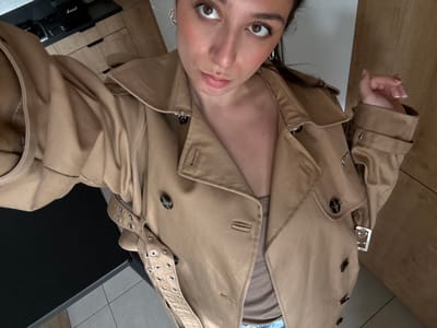 A female individual is depicted in a medium-shot, positioned at an overhead angle within an indoor residential setting. She wears a tan double-breasted trench coat with dark buttons and a decorative belt featuring metal eyelets, unfastened to reveal a light-colored top beneath. Her dark hair is pulled back, and she wears hoop earrings. Her right hand, exhibiting manicured nails with light polish, is visible near her left shoulder. The subject maintains direct eye contact with the camera, displaying a neutral facial expression. The background features light-toned wooden cabinetry, a dark countertop, and light-colored tiled flooring. A black amplifier, identifiable as a Marshall brand, is present on a higher shelf in the background. The specific location within the city of Blois, France, cannot be determined from the visual evidence provided.