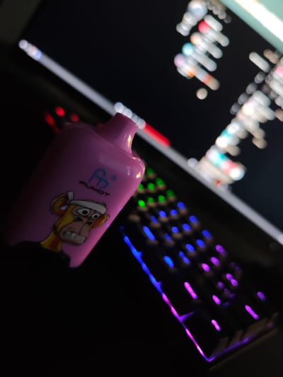 A close-up, dimly lit, and angled shot features a pink vape device with a cartoon ape graphic on its side. The vape rests on a keyboard with colorful LED backlighting. In the background, a computer monitor displays out-of-focus text and graphics, also illuminated by ambient light. The scene appears to be indoors, likely in a personal space given the setup. There are no people or animals present. The overall atmosphere is dark with points of vibrant color from the keyboard and monitor.