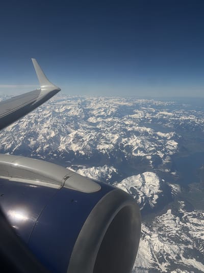 The image displays an aerial view from the window of an aircraft, capturing an expansive landscape of snow-capped mountain ranges under a clear, deep blue sky. In the upper left quadrant, the leading edge of a white aircraft wing, including an upward-curving winglet, is visible, with strong sunlight reflecting off its surface. Below it, occupying the lower-left to central foreground, is the forward section of a dark blue jet engine nacelle, featuring a prominent gray intake lip, also reflecting direct sunlight. The terrain below is characterized by numerous rugged mountains, with peaks and upper slopes heavily covered in white snow, interspersed with darker, exposed rock or vegetated areas. Towards the mid-ground on the right side, a large body of water, consistent with a lake, is discernible within the mountainous environment. The overall scene depicts a high-altitude flight over an alpine region. This scene is located in Peschiera Borromeo, Italy.