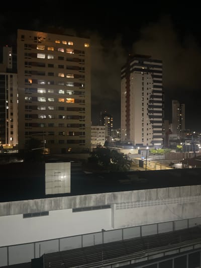 The image shows a nighttime cityscape in João Pessoa, Brazil, likely from an elevated perspective such as a hotel room or apartment balcony. Several multi-story buildings are visible, illuminated by interior lights and streetlights. The building on the left is a light-colored high-rise with numerous windows, many of which are lit. To its right, a taller building features a distinctive black and white striped facade, with scattered lit windows. Further in the background, other buildings of varying heights and designs are visible, some with lit windows. The sky is dark and cloudy, with some light reflecting from the city below. In the foreground, the rooftops of lower structures are visible, including what appears to be a building with a concrete facade and windows, and a darker corrugated roof. The overall scene depicts an urban environment at night, characterized by artificial lighting against a dark sky.
