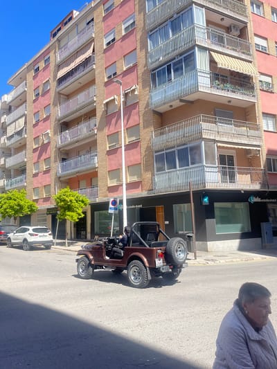 The image shows a street scene in Llíria, Spain, with a tall, multi-story apartment building on the left side of the frame. The building is primarily pink and beige, with white railings on the balconies. Several windows have awnings.

To the right of the building, on the ground floor, is a commercial space with a dark facade. A brown Jeep is parked on the street, partly obscuring the building's facade. A man is sitting in the driver's seat. A spare tire is attached to the back of the jeep.

Behind the jeep and towards the bottom of the frame, are parked cars and small green trees. A woman stands on the sidewalk in the upper right corner. The sky is clear blue.